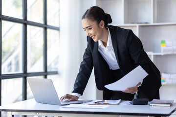 Young asian businesswoman sitting at desk with data and graph papers by using laptop at the office workplace, Accounting analyze, plan, and strategize about business improvement or finance.