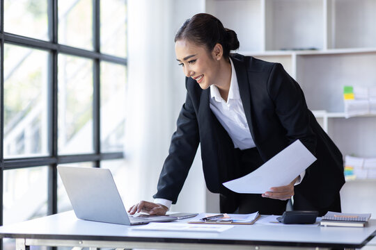 Young asian businesswoman sitting at desk with data and graph papers by using laptop at the office workplace, Accounting analyze, plan, and strategize about business improvement or finance.