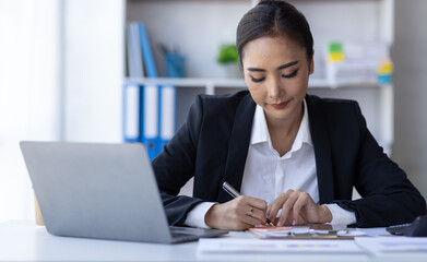 Young asian businesswoman sitting at desk with data and graph papers by using laptop at the office...