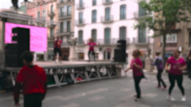 On stage, women show dance moves, passers-by repeat the movements. Performance in the city center. Background, blurry video.