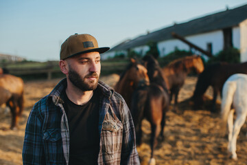 A young farmer near a herd of horses on a farm. Portrait of a bearded young man against the...