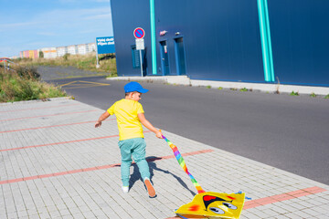 Little seven year old boy in a yellow T-shirt and blue baseball cap, white sneakers running with his yellow kite emoji outside against a blue wall