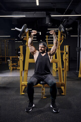 Tattooed, bearded male in black sweatpants, vest and cap. He performing a chest press while sitting...