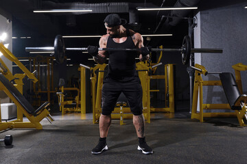 Bearded sportsman in black sport gloves, shorts, vest, cap and sneakers. He lifting a barbell, posing in dark gym with yellow equipment. Full length