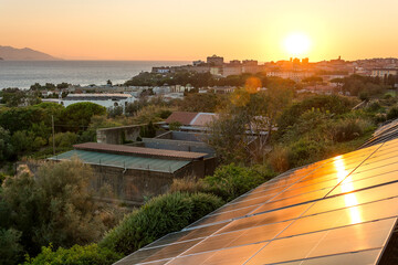 The setting sun is reflected in solar panels installed on a hillside with the city and the sea in...