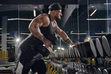 Young, tattooed, muscular man in black shorts, vest and cap. Exercising with dumbbells for training...
