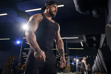Tattooed male in black shorts, vest, cap. Going to do exercises with dumbbells, looking at mirror near set of black weights. Dark gym. Close up