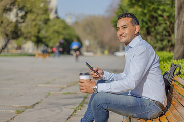 Portrait of latino man in shirt with a disposable cup of coffee sitting on a bench in a public park.