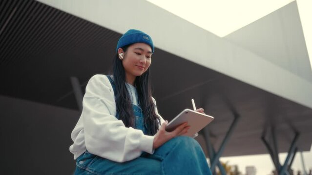 Cute Asian young woman in a hat with a tablet on the street. Drawing on a graphics tablet. Freelancing and earning online.
