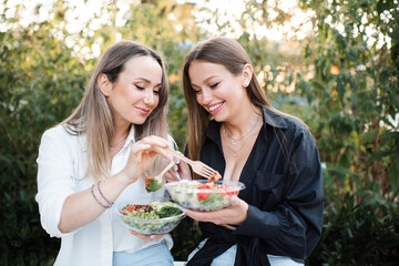 Two women friends eating fresh salads in bowls outdoor together. Friendship. Healthy eating