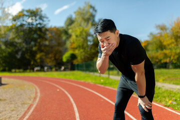 Asian young man doing sports at the stadium. He stands bent over on the treadmill, covers his mouth with his hand, feels very nauseous.