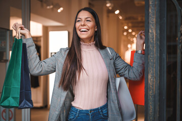 Attractive young woman feeling satisfied and joyful while walking out the clothing store in a shopping mall and holding colorful shopping paper bags with clothes.