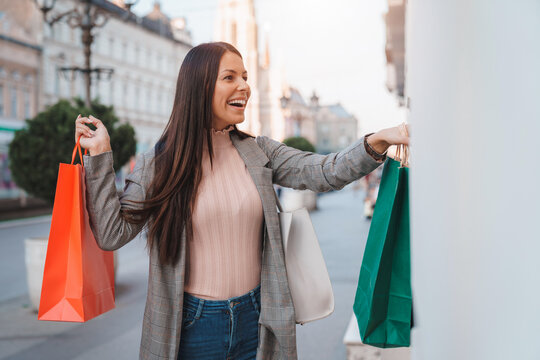 Attractive smiling cheerful woman with long hair holding paper shopping bags and pointing at store window while walking down the city street.
