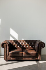 Vintage quilted classic brown sofa against white wall with sunlights from window on concrete floor at studio. Natural leather with rhombic stitching with buttons. Furniture design. Copy space