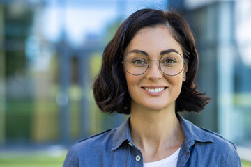 Close-up portrait of a young beautiful woman in glasses and a shirt, a student smiling and looking at the camera outside a university campus on a sunny day - Powered by Adobe