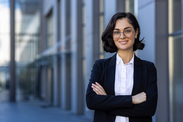 Young beautiful businesswoman in business suit smiling and looking at camera, portrait of successful woman outside office building, female worker in glasses with crossed arms