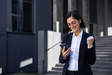 Young beautiful business woman in a business suit walking in the city with a phone in hands, a woman uses an application received a notification of a victory win, celebrates triumph and achievement