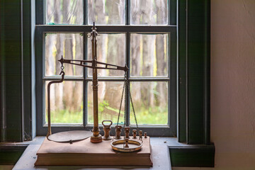 Antique Brass Balance Weight Scales in Front of Wavy Glass Multi Paned Wooden Window in Fort William Historical Park, Thunder Bay, Ontario, Canada