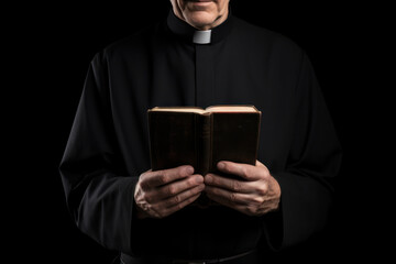 Catholic Priest in Black Cassock Holding Holy Bible: A Poignant Portrait Isolated on Black Background. - obrazy, fototapety, plakaty