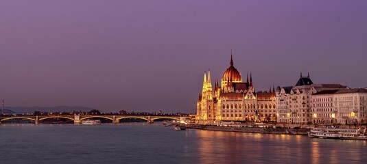 Fototapeta premium Panoramic view of illuminated grand buildings at the shore near a bridge at sunset