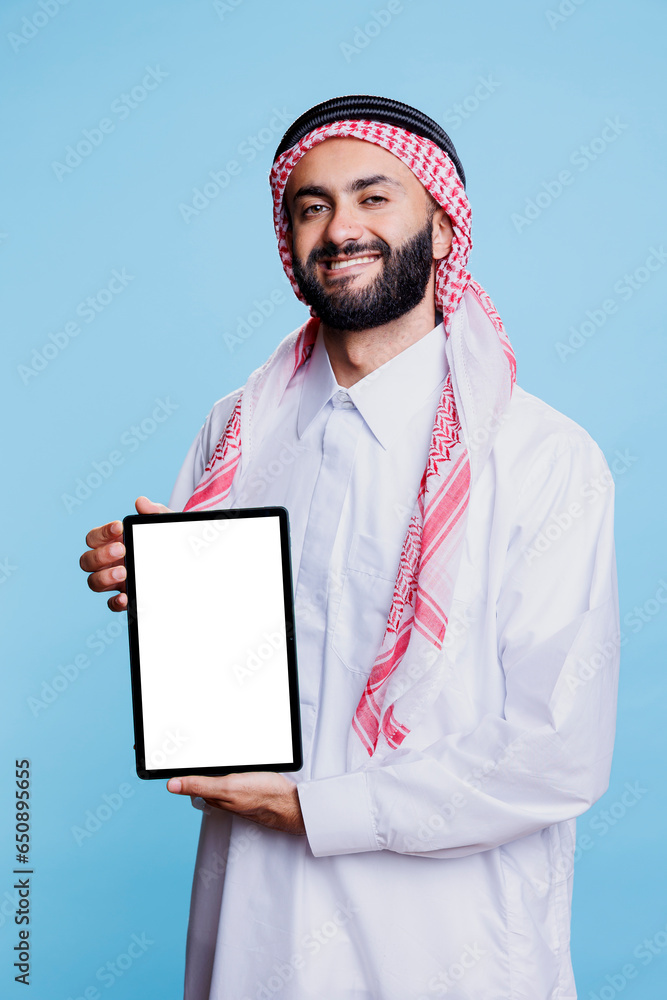 Poster smiling muslim man showing digital tablet with empty white screen and looking at camera with cheerfu