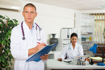 Positive male doctor standing in modern office with documents, writing medical history sheet