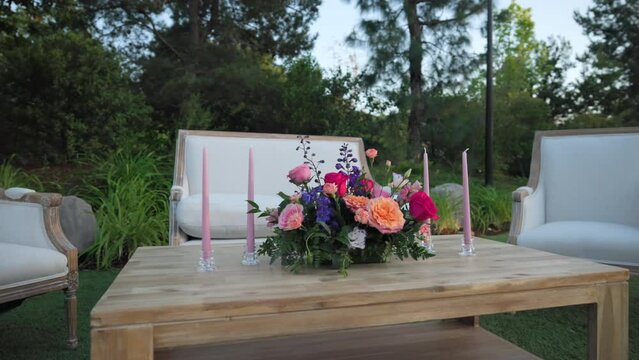 Small wooden table with colorful bouquet and four pink candles, white couch, two white armchairs standing in the green yard for outdoor wedding ceremony. High quality 4k footage