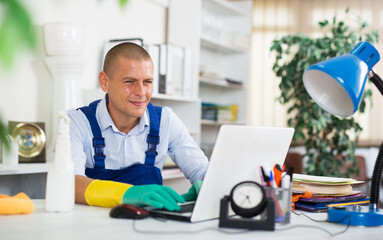 Smiling cleaning service worker in overalls and rubber gloves sitting at office table with laptop, chatting in social networks