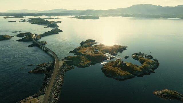 Drone flies over epic Atlantic Ocean road towards small island with houses in the middle of vast water surface in bright sunlight