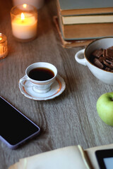 Vintage books, e-reader, pen, reading glasses, phone, cup of tea or coffee, bowl of cookies, green apple and lit candles on the table. Hygge at home, studying atmosphere. Selective focus.