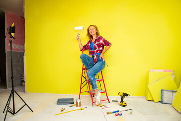 A beautiful young woman n a red plaid shirt, blue jeans and white sneakers  sits on a red stepladder with a roller in her hand against the background of a bright yellow wall - 650910474