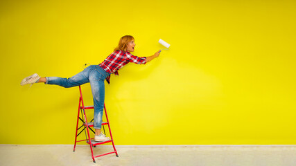 A beautiful young woman in a red plaid shirt, blue jeans and white sneakers  stands on a red stepladder and in an unusual pose with an outstretched leg stretches to paint the wall - 650910476