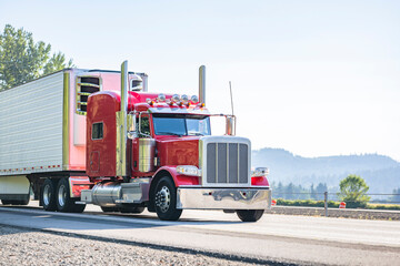 Bright red classic popular big rig semi truck tractor with chrome parts transporting frozen cargo in reefer semi trailer running on the highway road at sunshine