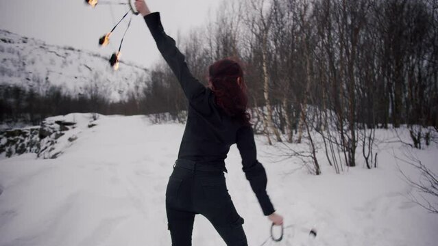 The beautiful young caucasian girl with red hair in a black shirt and black jeans masterly spins staff fire fans in the winter forest with snow, trees and hills. Perfomance with staff in a fire show