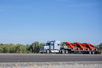 Light blue classic big rig semi truck tractor transporting equipment on step deck semi trailer...