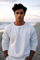 Young Hispanic man posing in a white crewneck, looking at the camera, against a softly focused beach shoreline.