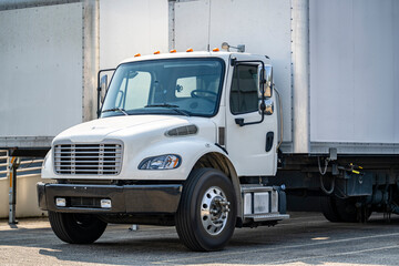 White compact middle duty day cab rig semi truck with box trailer loading cargo for the next delivery standing in warehouse dock with gates and another trailers