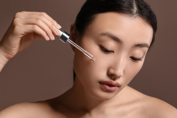 Fototapeta premium Beautiful young woman applying cosmetic serum onto her face on brown background, closeup