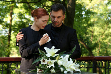 Sad couple mourning near granite tombstone with white lilies at cemetery. Funeral ceremony - obrazy, fototapety, plakaty