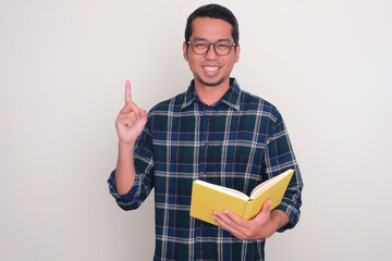 Adult Asian man smiling and pointing finger up while holding a book