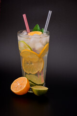 Tall faceted glass of refreshing lemonade with ice, straws and slices of orange and lemon on a black background.