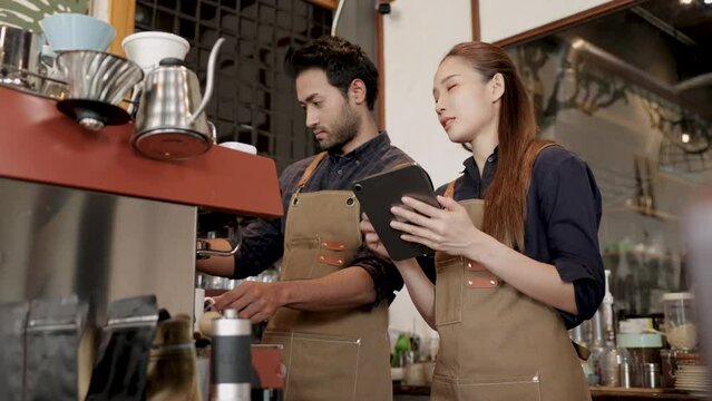 Cafe small business shop owner barista woman wearing apron call orders from tablet to coffee maker man, Indian husband and Asian wife shopkeeper partner run business start-up cafeteria and restaurant