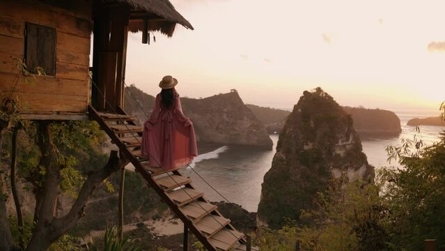Nusa penida, Bali, indonesia, 4K video of woman pink dress stand of tree house and looking at Atuh beach on tree house, Nusa Penida island at sunrise. Popular travel destination on Bali, indonesia.