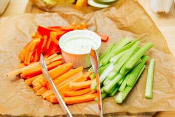 a wooden cutting board topped with sticks of carrots and celery with sauce on paper on the catering...