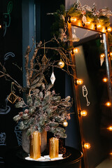 Tree branch decorated with hanging toys, standing besides candles and a mirror with warm bulbs. Interior in black and gold colors, holiday decoration.