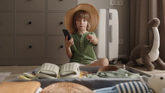 Little boy packs suitcase for travel. Serious, responsible, accountable, independent boy child kid with smartphone in hands and checklist, checks presence of all clothes in suitcase baggage luggage. 