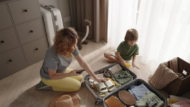 Mother and son packing suitcase together. Mom with smartphone checks checklist, packing suitcase with son in organized attentive manner. Mom kid give high five congratulating on end of packing baggage