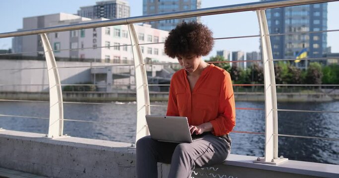 young african american woman sitting at embankment with laptop online typing working remotely