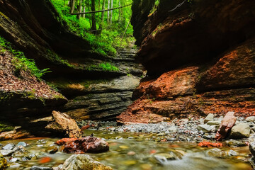 entry to a narrow canyon with layers of red rocks and a brook under a forest