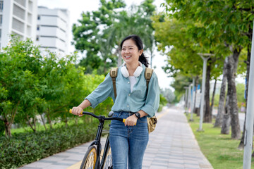 Young Asian woman walks side by side with a bicycle in summer in a park on a city street.